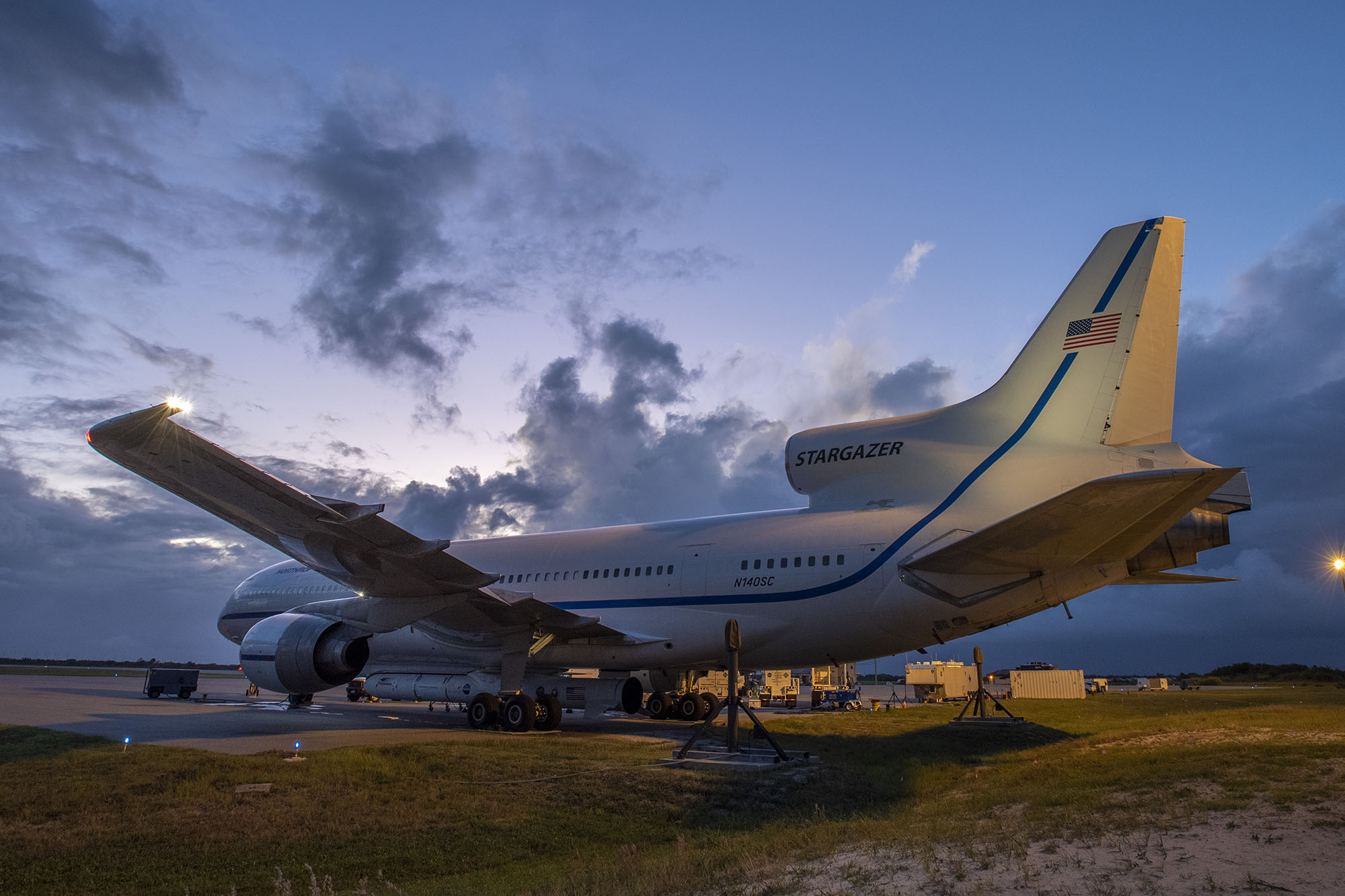 Rear side view of airplane parked off the side of a runway in front of dark blue sky at sunset